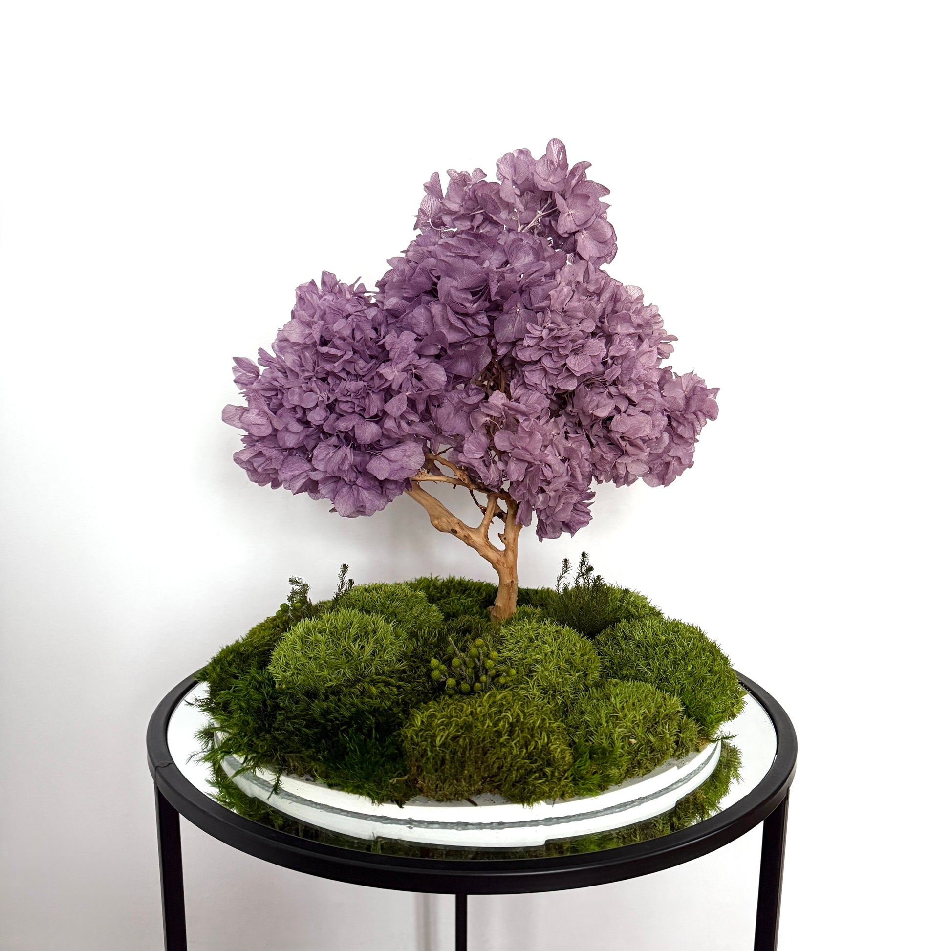 Flowering bonsai zen garden with lavender hydrangea canopy and layered moss landscape on a round white base, styled as Japandi console decor.